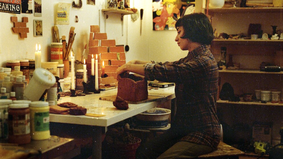Lizette Hernández is seated in front of a table, sculpting a container out of clay, using her hands. She is wearing a button-up plaid shirt and her hair is cut short in a bob. She's surrounded by lit candle sticks on the table and art materials and projects in various stages of completion. 