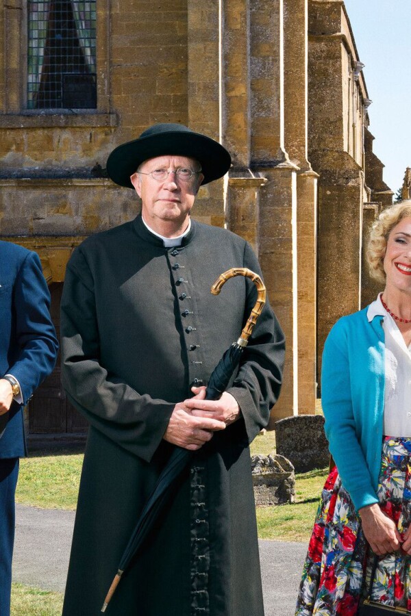Five members of the cast of "Father Brown" pose side by side beside a small road and a vintage car.