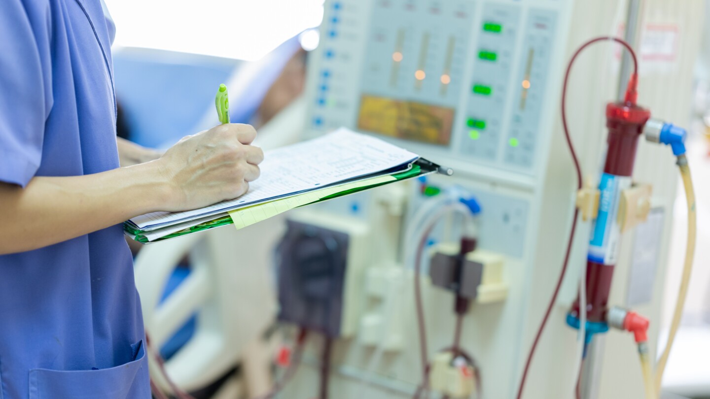 A technician wearing blue scrubs holds a clipboard as they check continuous renal replacement therapy equipment and injection pump and hemodialysis machine.