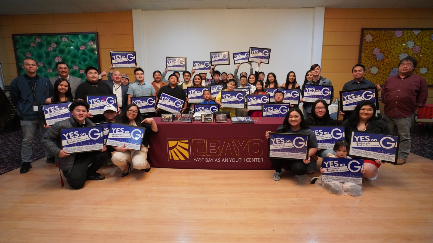 Young Asian Americans hold up signs that say "Yes on G" with a table that says EBAYC (East Bay Asian Youth Center)