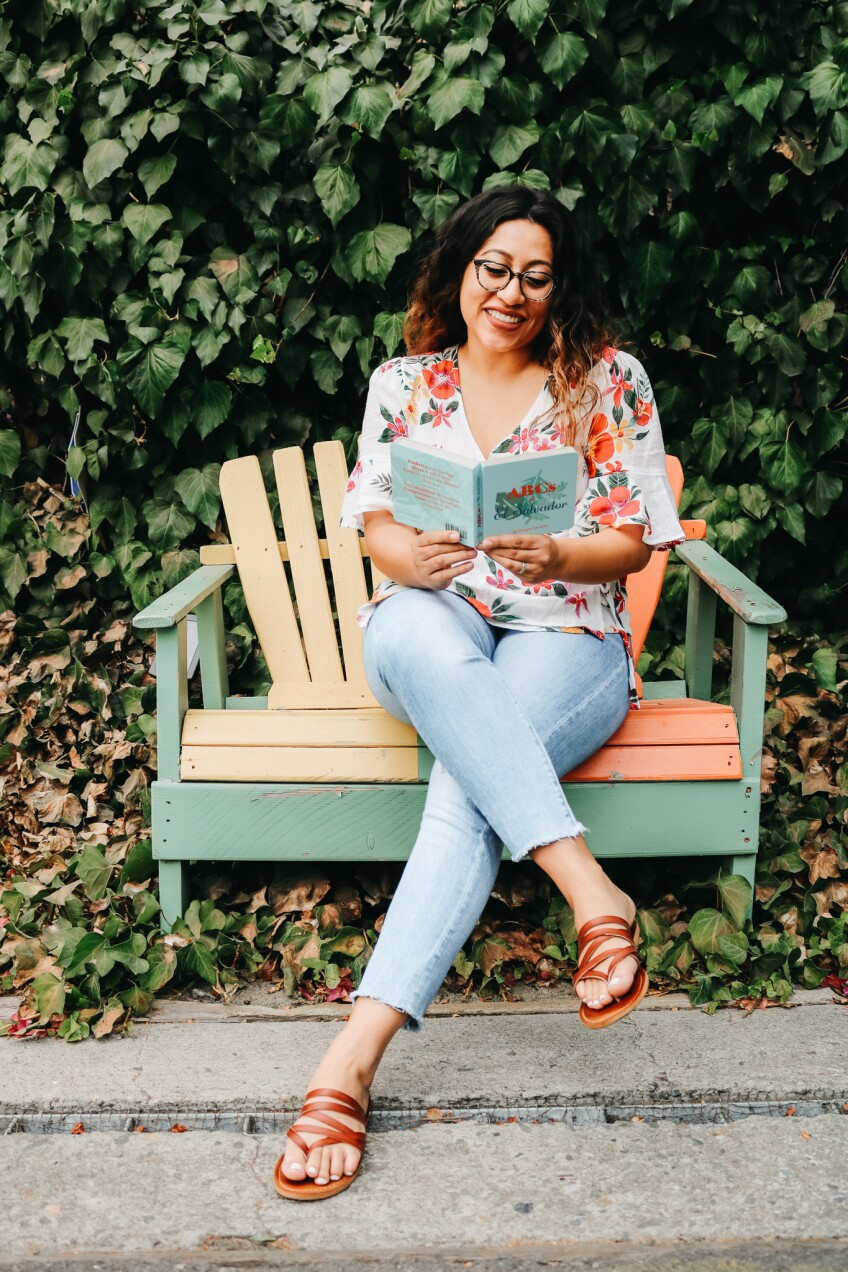 Author Maggie Carranza poses on a chair outside with her book "ABCs of El Salvador"