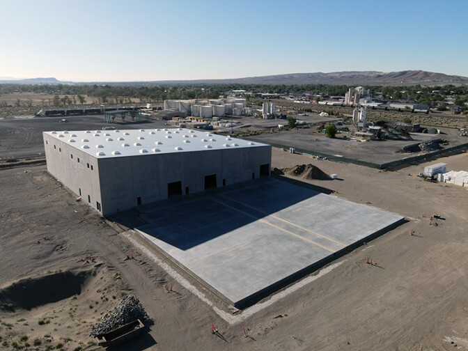 A concrete slab juts out of a rectangular plant building in a large dirt lot with mountains in the distance.