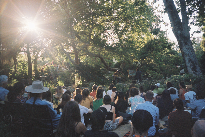A crowd of audience members are seated near each other on the ground at a Japanese Garden, surrounded by tall, winding trees that tower above them. To the top left corner, sun rays filter through the leaves, creating a starburst in the photo. A three-piece music ensemble is seen in the background, performing on a small make-shift stage.