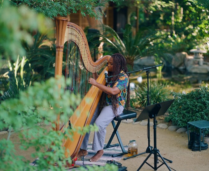 A musician wearing a bright, colorful, floral printed top is seated while playing a large wooden harp that is resting on their shoulder and thigh. There is a microphone stand with a microphone placed at their lips. They are playing outdoors, amongst bright green foliage. 