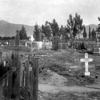 A black and white archival image of a cemetery with gravesites sparsely scattered across the ground, marked various sized and shaped crosses. Many of the crosses are encircled with a wooden fence. Beyond, is a mountain and trees. 