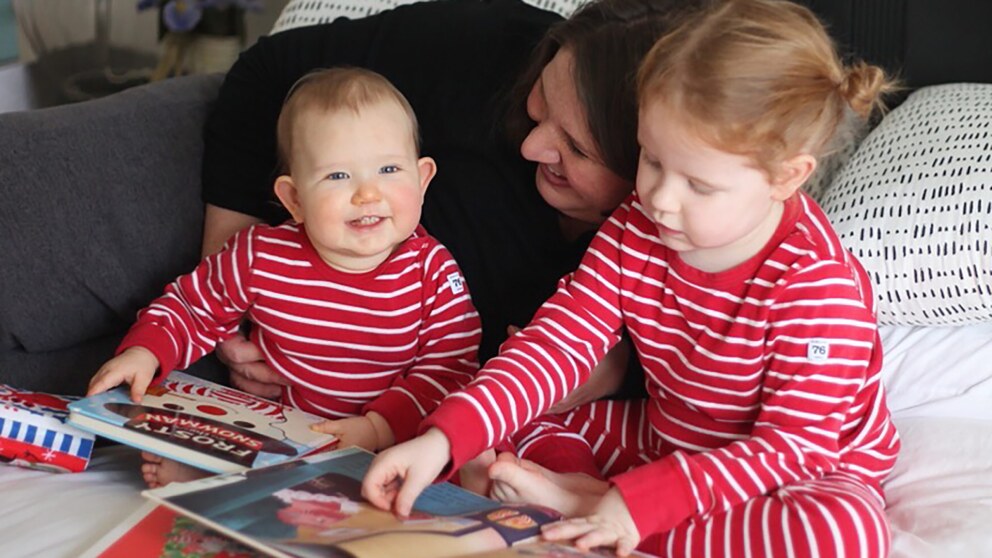 A woman cuddles two small children in matching pijamas as they read together on a bed.