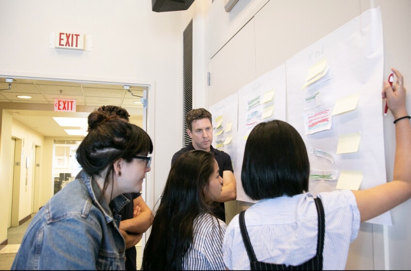 A group of students gather around a wall covered in sticky notes. 