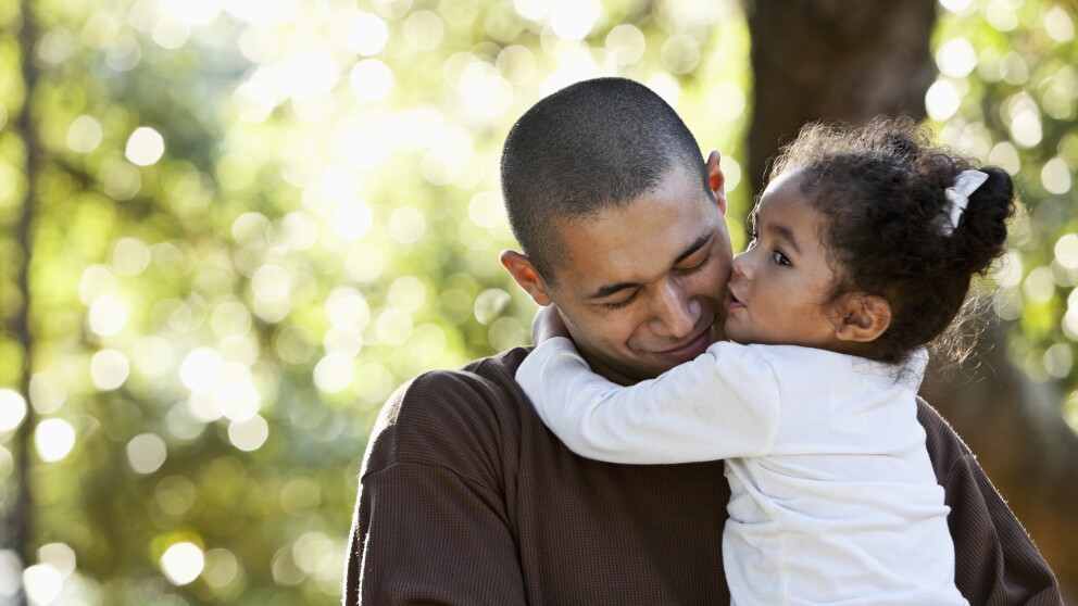 An Hispanic father holding his daughter in his arms on a sunny day at the park.  The beautiful little girl, 2 years old, has her arms around daddy's neck and is kissing him on the cheek.  He is smiling with his eyes closed.