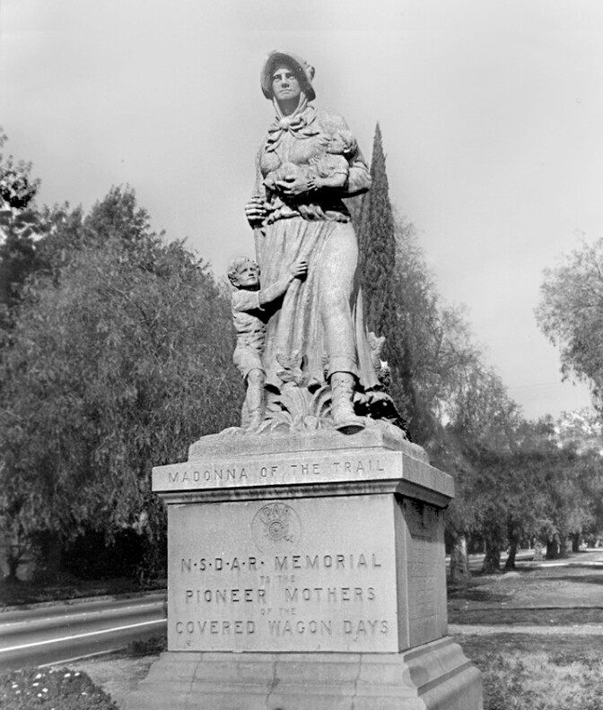A black and white photo of a stone sculpture of a woman, dressed in a attire that old Californian pioneers would wear. She looks sternly off into the distance while cradling a baby in one arm and a rifle in the other. A young boy clutches on her leg, tugging at the 