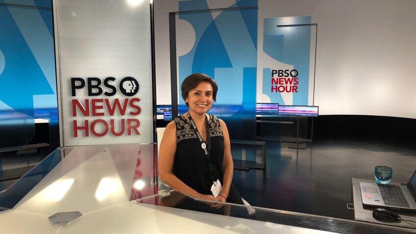 A woman poses at a desk in a tv news set.
