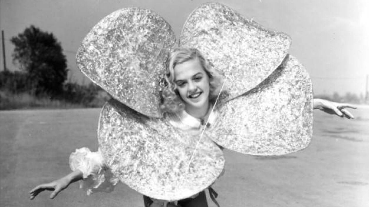 A vertical, black and white portrait of a blonde woman wearing a sparkly four-leaf clover costume as she holds her arms out and extends a leg as though in a curtsy.